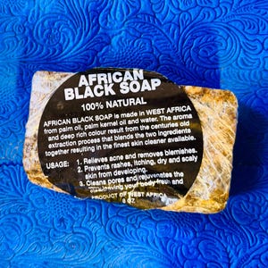 Puede incluir: Una barra de jabón negro africano, envuelta en plástico transparente, con una etiqueta negra. La etiqueta dice "African Black Soap 100% Natural". El jabón está hecho en África Occidental con aceite de palma, aceite de palmiste y agua. Pesa 227 gramos.