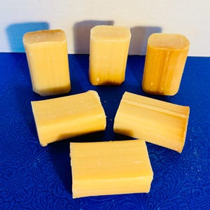 Puede incluir: Cinco barras rectangulares de jabón amarillo dispuestas sobre una superficie azul. Las barras de jabón tienen una forma ligeramente redondeada y una textura suave. La iluminación resalta las variaciones naturales de color del jabón.