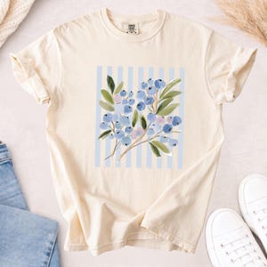 May include: A cream-colored short-sleeve t-shirt featuring a central graphic of blue berries and green leaves layered over a light blue and white vertical striped background. The shirt has a crew neckline and a standard fit with rolled sleeves.