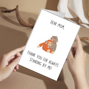 May include: A greeting card with the text "Dear Mom, Thank you for always standing by me!" features an illustration of two monkeys hugging. The card is held by hands against a neutral background.