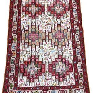 Puede incluir: Una alfombra rectangular con un campo central blanco y un borde rojo. La alfombra presenta un patrón geométrico repetitivo en rojo, beige y otros colores. El borde tiene diseños florales y geométricos. La alfombra tiene flecos en los extremos cortos.