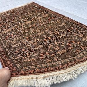 Könnte beinhalten: Ein rechteckiger, handgewebter Teppich mit einem detaillierten geometrischen Muster in Braun-, Rost- und Beigetönen. Der Teppich hat einen Fransenrand und scheint aus Naturfasern hergestellt zu sein. Das Design weist sich wiederholende Motive auf.