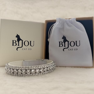 Könnte beinhalten: Ein weißes Katzenhalsband mit zwei Reihen funkelnder, klarer Kristalle. Das Bild zeigt das Halsband zusammen mit einer weißen Schachtel und einem Kordelzugbeutel, beide mit dem Logo "Bijou Cat Co" und einer Katzen-Silhouette.