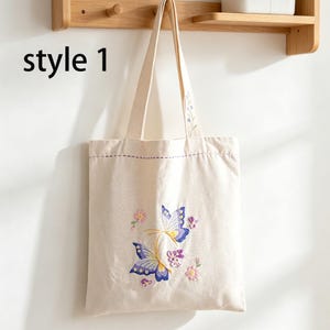 May include: A cream-colored tote bag with embroidered butterflies and flowers. The bag has a rectangular shape with a purple stitched border and long straps. The text "style 1" is in the upper left corner.