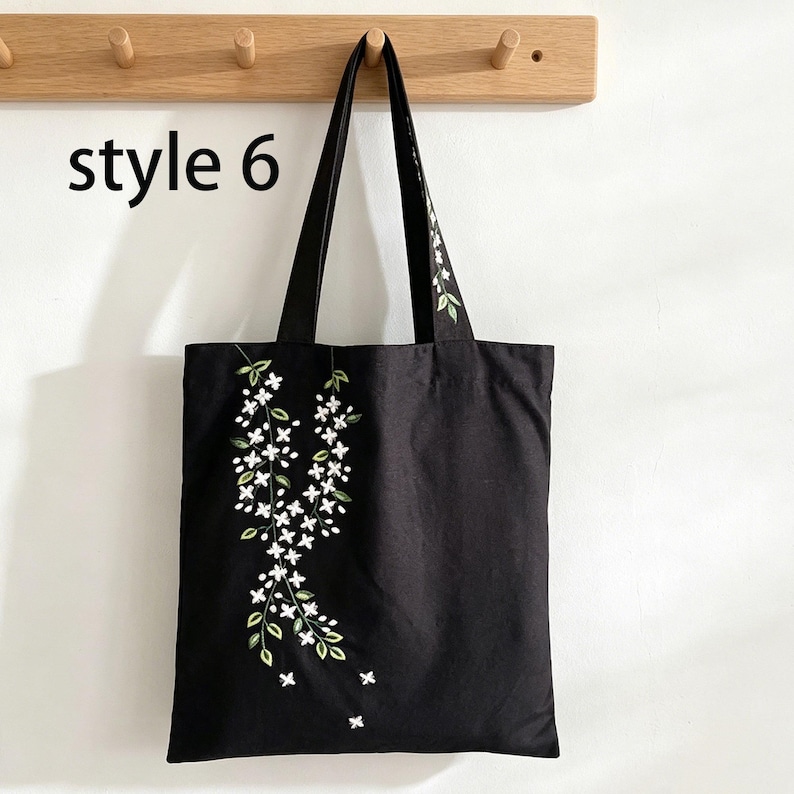 May include: Black tote bag with white floral embroidery and green leaves. The bag has long handles and the text "style 6" is visible on the upper left. The bag is hanging on a wooden rack.
