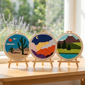 Könnte beinhalten: Drei runde Stickrahmen mit Landschaftsmotiven. Einer zeigt eine Wüstenlandschaft mit Kaktus und Sonne, ein anderer eine Bergkette mit Mond, und der dritte eine Berglandschaft mit Bäumen und einem See. Jeder wird auf einem kleinen Holzstaffelei präsentiert.