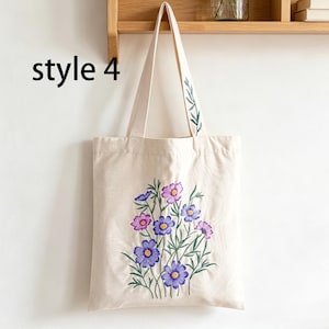 May include: Beige canvas tote bag with embroidered purple and pink flowers and green leaves. The bag has long handles and the text "style 4" in black on the left side. The bag is hanging on a wooden shelf.