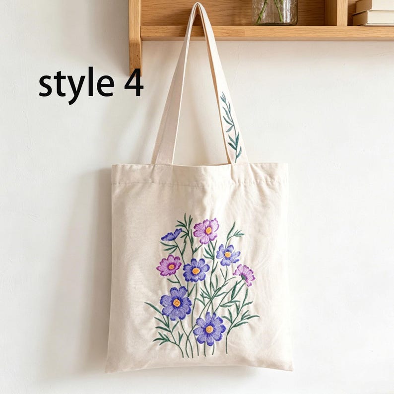 May include: Beige canvas tote bag with embroidered purple and pink flowers and green leaves. The bag has long handles and the text "style 4" in black on the left side. The bag is hanging on a wooden shelf.
