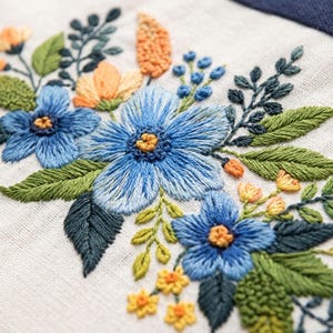 May include: Close-up of a floral embroidery design featuring blue flowers with yellow centers, orange accents, and green leaves. The embroidery is on a white fabric background, showcasing detailed stitching.