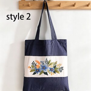 May include: A navy blue tote bag with a white horizontal stripe featuring an embroidered floral design. The flowers are blue, orange, and yellow with green leaves. The text "style 2" is in the upper left corner.