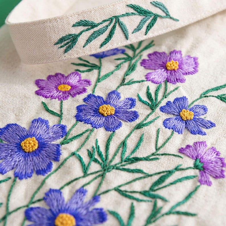 May include: Close-up of embroidered flowers in shades of purple and blue with yellow centers on a cream-colored fabric. Green leaves and stems are also embroidered. A cream-colored band with green embroidered leaves is at the top.