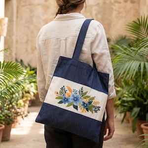 May include: A denim tote bag with a cream-colored stripe featuring an embroidered floral design in blue, orange, and green. The bag has a dark blue handle and is carried over the shoulder.