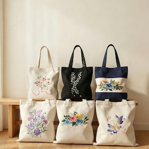 May include: Six canvas tote bags with embroidered floral designs. The bags are in various colors, including cream, black, and navy blue. The floral patterns feature a variety of colors, including pink, blue, yellow, and purple. The bags have long handles.