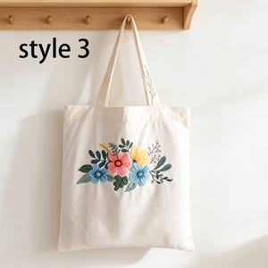 May include: Cream-colored tote bag with embroidered floral design. The flowers are in shades of pink, blue, and yellow, with green leaves. The bag has a simple design and the text "style 3" is printed on the upper left.