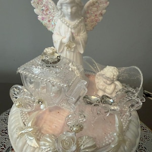 May include: A white ceramic angel figurine with glitter-covered wings. The angel stands above a decorative base with a carriage, cherub, and floral details. The base has a pink centre and pearl embellishments. The base is white.