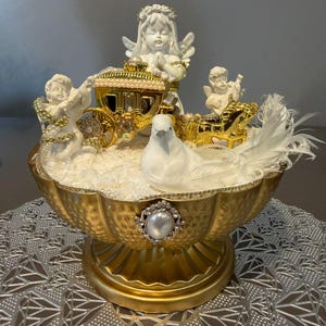 May include: A decorative gold-coloured centrepiece featuring cherubic figurines, a carriage, and a white dove. The centrepiece has a scalloped bowl shape with a pearl and rhinestone embellishment. White feathers add a delicate touch.