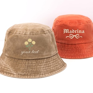 May include: A brown bucket hat featuring an embroidered yellow flower design and the text "your text" alongside an orange bucket hat with the embroidered text "Madrina" and a white bow graphic. Both hats have a washed, textured fabric finish.
