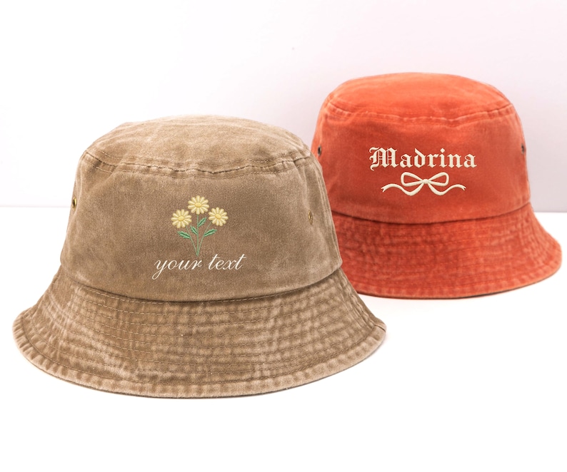 May include: A brown bucket hat featuring an embroidered yellow flower design and the text "your text" alongside an orange bucket hat with the embroidered text "Madrina" and a white bow graphic. Both hats have a washed, textured fabric finish.