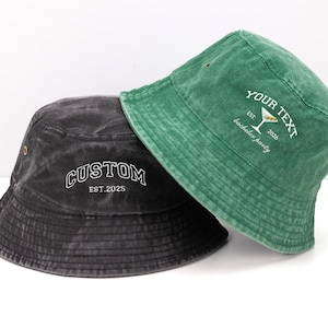 May include: A charcoal gray bucket hat with the white embroidered text CUSTOM EST.2025 and a green bucket hat with white embroidered text reading YOUR TEXT EST. 2026 bachelor party featuring a martini glass graphic with two olives.