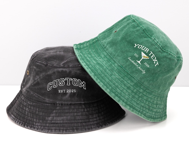 May include: A charcoal gray bucket hat with the white embroidered text CUSTOM EST.2025 and a green bucket hat with white embroidered text reading YOUR TEXT EST. 2026 bachelor party featuring a martini glass graphic with two olives.