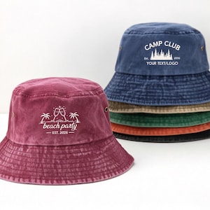May include: A stack of five cotton bucket hats in maroon, blue, tan, green, orange, and black. The top blue hat features white text reading CAMP CLUB EST. 2026 YOUR TEXT/LOGO. The front maroon hat displays white text reading beach party EST. 2026.