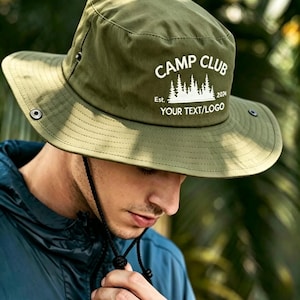 Könnte beinhalten: Ein olivgrüner Fischerhut mit breiter Krempe und schwarzem Kinnriemen. Auf der Vorderseite befindet sich ein weißer Schriftzug mit CAMP CLUB, Est. 2024 und YOUR TEXT/LOGO sowie eine grafische Darstellung eines Kiefernwaldes.
