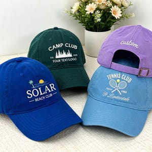 May include: A collection of four baseball caps in blue, dark green, light blue, and purple. The blue cap features a yellow sun and palm tree graphic with the text SOLAR BEACH CLUB. The green cap displays a forest graphic and CAMP CLUB text.