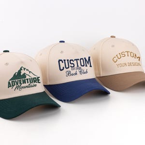 Könnte beinhalten: Drei Baseballkappen mit gestickten Designs. Eine Kappe ist beige mit grünem Schirm und einer Berg-Grafik mit dem Text "Adventure Mountain". Eine andere ist beige mit blauem Schirm und dem gestickten Wort "CUSTOM". Die dritte ist beige mit braunem Schirm und dem gestickten Wort "CUSTOM".