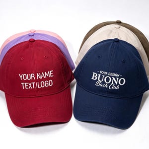 May include: A collection of baseball caps in various colors including red, purple, pink, navy blue, beige, and brown. The red cap features the white text "YOUR NAME TEXT/LOGO" and the navy blue cap features the white text "YOUR DESIGN BUONO Bach Club".