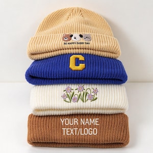 May include: A stack of four ribbed knit beanies. The top beige beanie features embroidered animal faces and the text BE HAPPY EVERY DAY. The blue beanie has a yellow letter C. The white beanie has purple flowers. The brown beanie has YOUR NAME TEXT/LOGO.