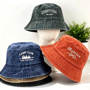 May include: Three cotton bucket hats in charcoal, navy blue, and burnt orange. The charcoal hat features white text reading CUSTOM EST.2025. The navy hat displays white text reading CAMP CLUB EST. 2016 YOUR TEXT/LOGO. The orange hat features white text reading Madrina.