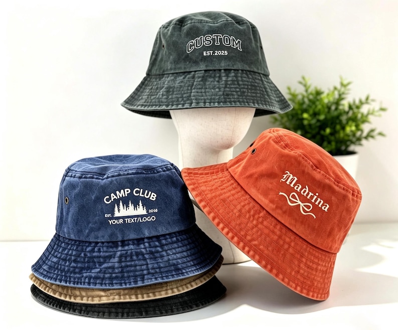 May include: Three cotton bucket hats in charcoal, navy blue, and burnt orange. The charcoal hat features white text reading CUSTOM EST.2025. The navy hat displays white text reading CAMP CLUB EST. 2016 YOUR TEXT/LOGO. The orange hat features white text reading Madrina.
