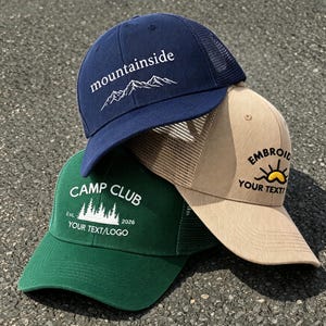 Pode incluir: Três bonés trucker em azul marinho, verde floresta e bege. O boné azul apresenta gráficos de montanhas brancos e o texto "mountainside". O verde exibe "CAMP CLUB" com gráficos de floresta e "YOUR TEXT/LOGO". O bege mostra um ícone de sol e "EMBROID YOUR TEXT".