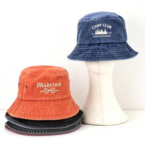 May include: A stack of four bucket hats in orange, dark gray, and blue. The top blue hat features white text reading CAMP CLUB Est. 1926 YOUR TEXT/LOGO with a forest graphic. The orange hat features the white text Madrina with a bow graphic.