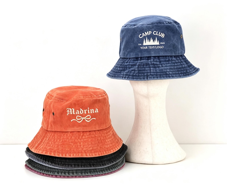 May include: A stack of four bucket hats in orange, dark gray, and blue. The top blue hat features white text reading CAMP CLUB Est. 1926 YOUR TEXT/LOGO with a forest graphic. The orange hat features the white text Madrina with a bow graphic.