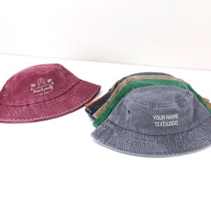 May include: A collection of cotton bucket hats in various colors including maroon, blue, green, and tan. The maroon hat features a white graphic with two palm trees and the text beach party EST. 2020. The blue hat displays the text YOUR NAME TEXT/LOGO.