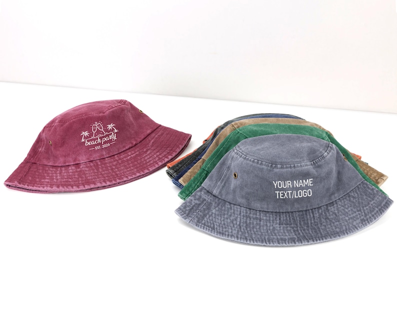 May include: A collection of cotton bucket hats in various colors including maroon, blue, green, and tan. The maroon hat features a white graphic with two palm trees and the text beach party EST. 2020. The blue hat displays the text YOUR NAME TEXT/LOGO.