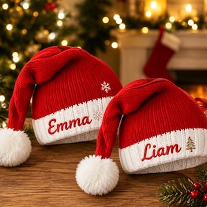 Könnte beinhalten: Zwei rot-weiße Strick-Weihnachtsmützen mit weißen Bommeln. Die linke Mütze ist mit dem Namen Emma und einer Schneeflocke bestickt. Die rechte Mütze trägt den Namen Liam und einen kleinen Weihnachtsbaum. Beide haben einen gerippten weißen Saum.