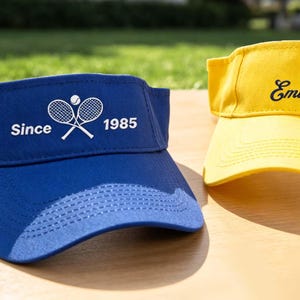 May include: A royal blue visor featuring a white graphic of two crossed tennis rackets, a tennis ball, and the text "Since 1985" alongside a yellow visor with a graphic of a basketball and the word "Embroidery" in black script lettering.