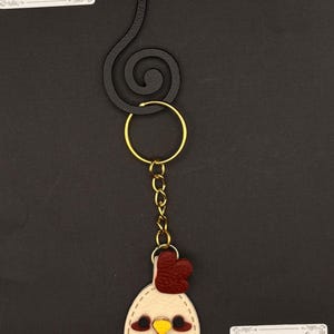 May include: A keychain featuring a cream-colored chicken head with a red comb and yellow beak. The chicken charm is attached to a gold-colored chain and ring, connected to a black metal swirl. The text "Moss & Ember Studio" is on a white label.