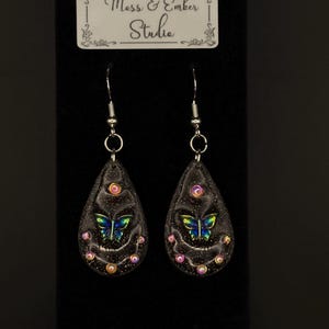Op de afbeelding: Een paar zwarte druppelvormige oorbellen met een vlinderontwerp. De oorbellen hebben zilveren haken en zijn versierd met iriserende glitter en kleine roze en gouden kralen. De tekst "Mess & Ember Studio" staat op een wit label.