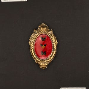 Puede incluir: Broche ovalado dorado con centro rojo y tres diseños de mariposas negras y amarillas. La broche está sobre un fondo negro. El texto "Mess & Ember Studio" es visible.