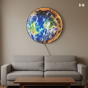 Op de afbeelding: Een ronde wandkunst die de aarde afbeeldt, met groene en bruine continenten, blauwe oceanen en een gloeiende oranje rand. Het kunstwerk hangt boven een grijze bank en een houten salontafel. De tekst "B-16" is zichtbaar in de hoek.