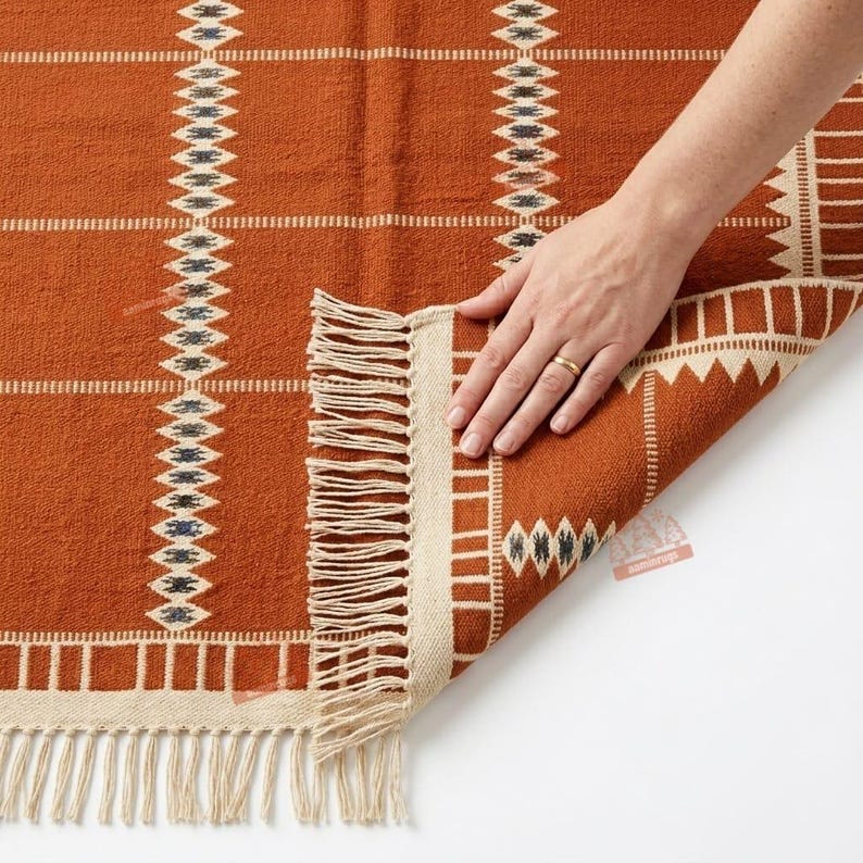May include: A woven rug in a deep burnt orange colour featuring a geometric pattern of white and dark blue diamonds arranged in vertical stripes. The rug has a cream-coloured border and a fringe edge along the bottom and side, with a textured weave.