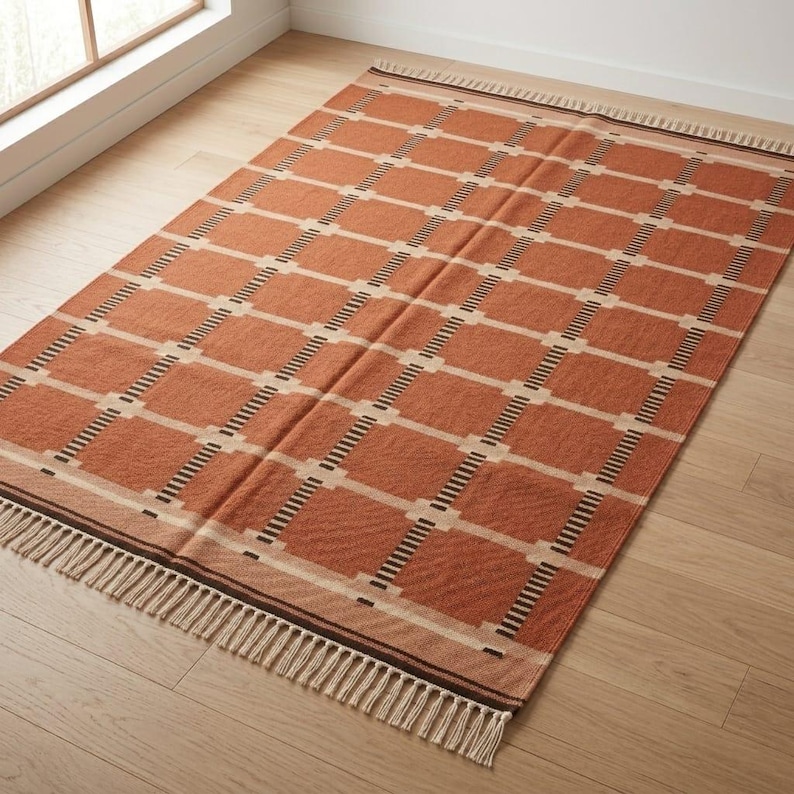May include: A rectangular area rug featuring a terracotta orange base with a repeating grid pattern of cream-colored lines and dark brown dashed accents. The rug is finished with short, light-colored fringe along the two shorter ends.