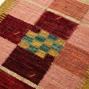 May include: A woven textile featuring a grid pattern of large squares in shades of deep red, dusty pink, and muted orange. A central square block displays a smaller, intricate checkerboard design in alternating teal and mustard yellow tones.