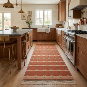 May include: A long rectangular runner rug featuring a terracotta orange base with a repeating grid pattern of cream-colored lines and small black accents, finished with a short cream-colored fringe along both of the shorter ends.