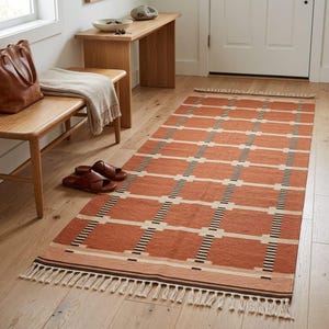 May include: A rectangular area rug featuring a terracotta orange base with a grid pattern of white horizontal and vertical lines accented by thin black stripes. The rug has a flat-woven texture and is finished with short fringe on both ends.