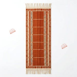 May include: A rectangular runner rug in a deep burnt orange colour featuring a cream-coloured geometric border pattern with diamond shapes and horizontal lines. The rug is finished with cream-coloured fringe tassels on both short ends.