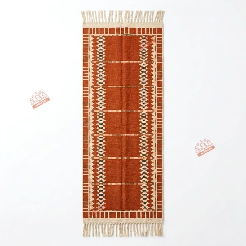 May include: A rectangular runner rug in a deep burnt orange colour featuring a cream-coloured geometric border pattern with diamond shapes and horizontal lines. The rug is finished with cream-coloured fringe tassels on both short ends.